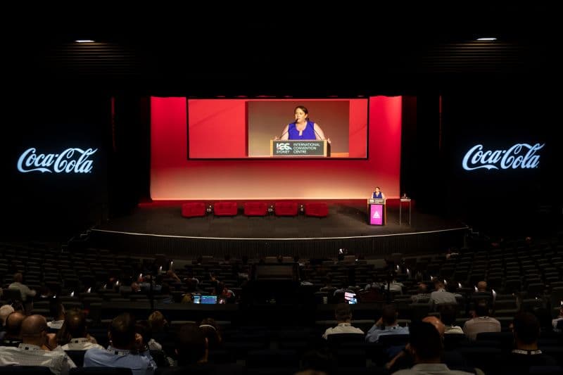 Bec presenting on a large conference stage between Coca-Cola screens