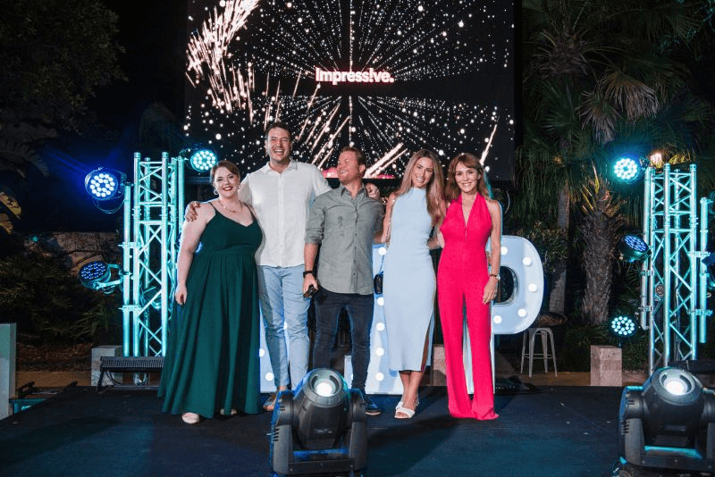 Five people standing together on a brightly lit awards stage, with VIP lettering and a fireworks display on the screen behind them.